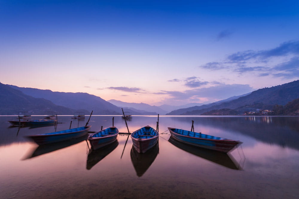 Vier Boote im ruhigen Wasser eines Sees bei Sonnenuntergang, umgeben von Bergen.