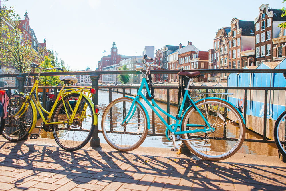 Zwei Fahrräder, eines gelb und eines türkis, stehen an einem Kanal in Amsterdam. Im Hintergrund sind historische Gebäude zu sehen.