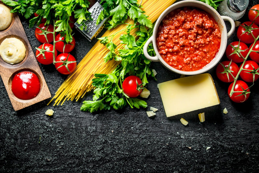 Zutaten für Spaghetti: frische Tomaten, Petersilie, Spaghetti, Hackfleischsauce und Käse auf einem dunklen Hintergrund.