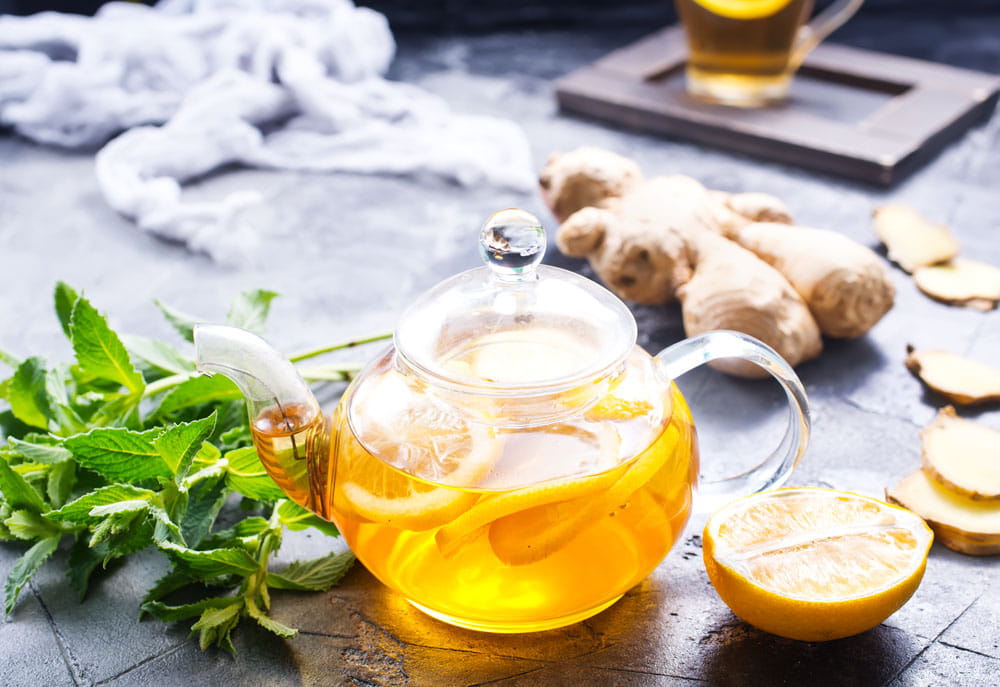 Teekanne mit Spritzschutz, gefüllt mit Ingwer-Zitronentee, frische Minze und Ingwerstücken im Hintergrund.