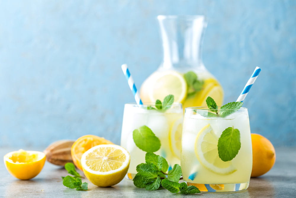 Zwei Gläser Zitronenlimonade mit Minze und Zitronenscheiben, im Hintergrund eine Karaffe mit Limonade und frischen Zitronen.