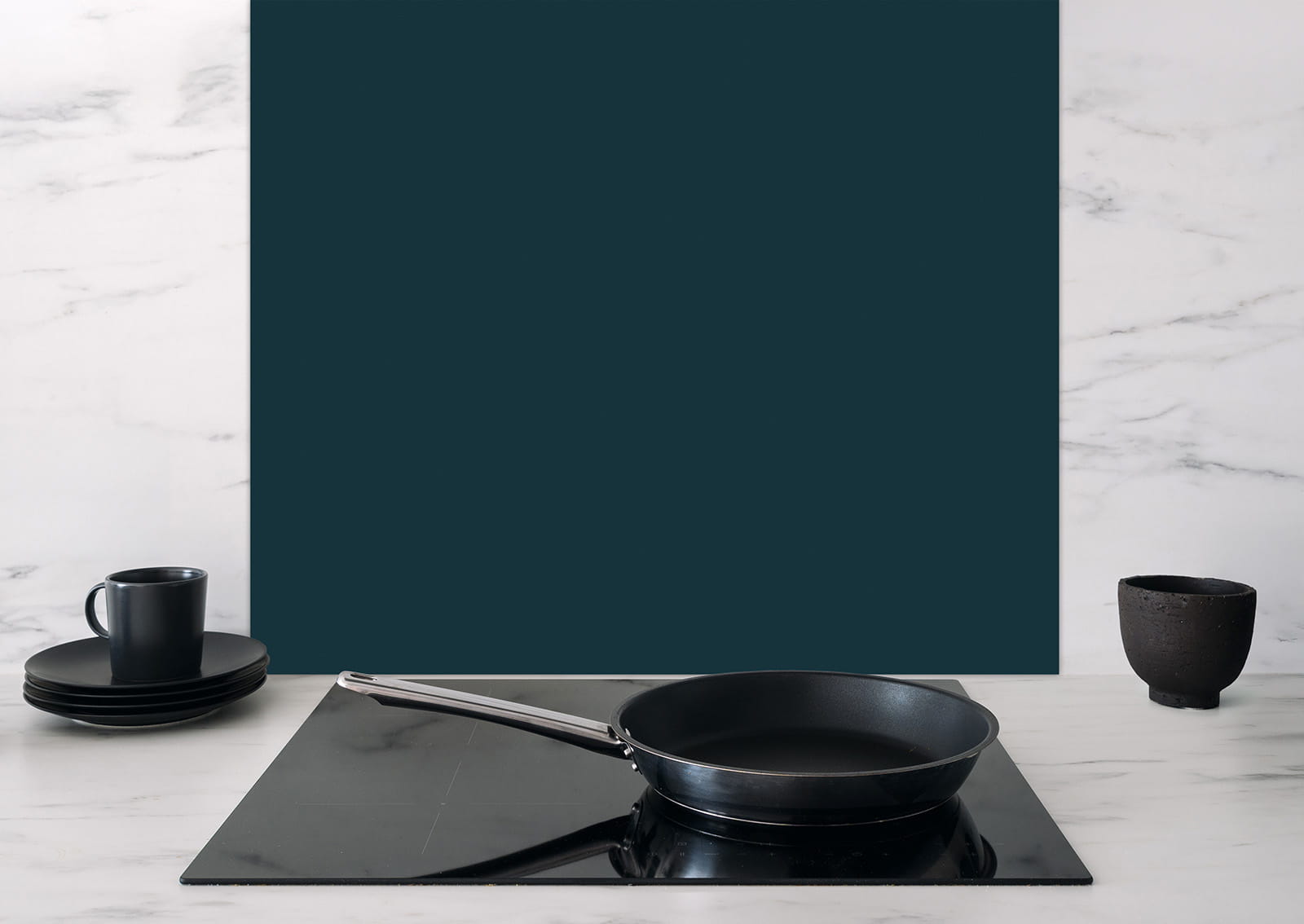 Spritzschutz Blaugrau von ENTDECKER hinter einer schwarzen Pfanne auf einem Kochfeld, mit Geschirr in Schwarz.